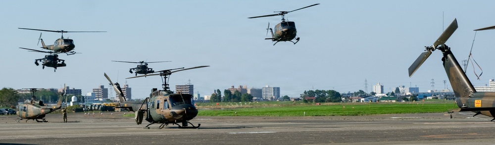 A military airfield scene showing multiple olive-drab/camouflage helicopters in various stages of takeoff, hovering, and ground positioning. At least six helicopters are visible, with several airborne and others on the tarmac. A urban skyline with buildings and trees is visible in the background.