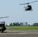 A military airfield scene showing multiple olive-drab/camouflage helicopters in various stages of takeoff, hovering, and ground positioning. At least six helicopters are visible, with several airborne and others on the tarmac. A urban skyline with buildings and trees is visible in the background.