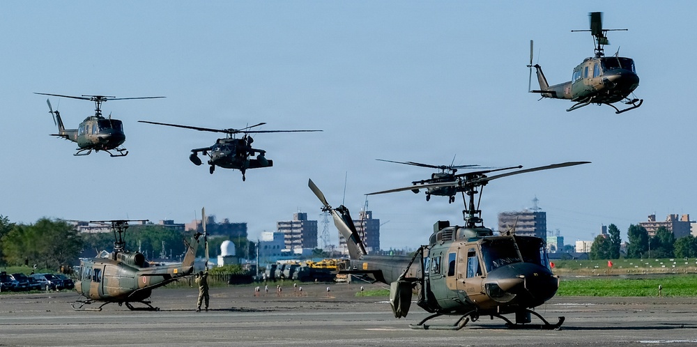 A military airfield scene showing multiple helicopters in various stages of takeoff, landing, and ground operations. At least six helicopters are visible, including what appear to be UH-1 Iroquois (Huey) variants and a Black Hawk-type helicopter, consistent with Japan Ground Self-Defense Force markings and olive drab color schemes. Urban buildings are visible in the background, suggesting proximity to a populated area.