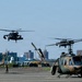A military airfield scene showing multiple helicopters in various stages of takeoff, landing, and ground operations. At least six helicopters are visible, including what appear to be UH-1 Iroquois (Huey) variants and a Black Hawk-type helicopter, consistent with Japan Ground Self-Defense Force markings and olive drab color schemes. Urban buildings are visible in the background, suggesting proximity to a populated area.