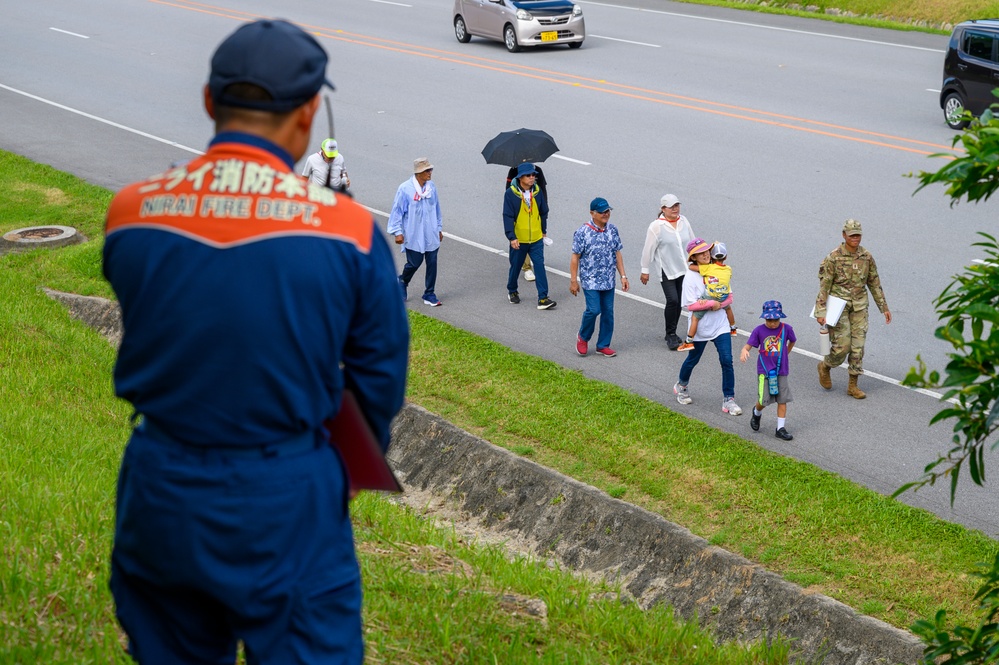 A Nirai Fire Department official in a blue uniform observes a group of civilians and a military personnel walking along a road shoulder. The scene appears to be in Okinawa, Japan, based on the Japanese text on the official's uniform and the local road markings. A mix of adults and children are walking together, with one person carrying an umbrella and another holding a child.