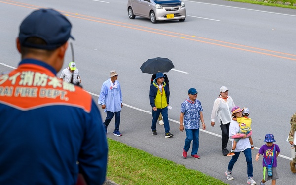 A Nirai Fire Department official in a blue uniform observes a group of civilians and a military personnel walking along a road shoulder. The scene appears to be in Okinawa, Japan, based on the Japanese text on the official's uniform and the local road markings. A mix of adults and children are walking together, with one person carrying an umbrella and another holding a child.