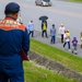 A Nirai Fire Department official in a blue uniform observes a group of civilians and a military personnel walking along a road shoulder. The scene appears to be in Okinawa, Japan, based on the Japanese text on the official's uniform and the local road markings. A mix of adults and children are walking together, with one person carrying an umbrella and another holding a child.