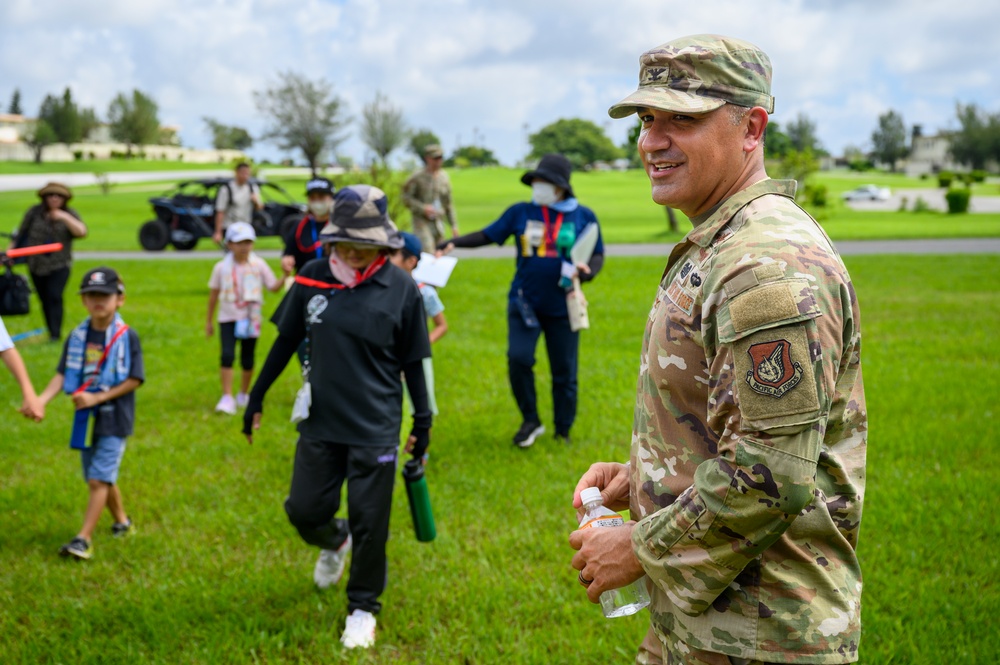 A U.S. Air Force service member in OCP (Operational Camouflage Pattern) uniform stands smiling on a grassy field while a group of children and adults gather behind him. The patch on his shoulder indicates affiliation with the U.S. Air Force. The setting appears to be an outdoor community or educational event, possibly on or near a military installation.