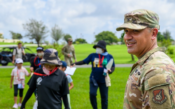 A U.S. Air Force service member in OCP (Operational Camouflage Pattern) uniform stands smiling on a grassy field while a group of children and adults gather behind him. The patch on his shoulder indicates affiliation with the U.S. Air Force. The setting appears to be an outdoor community or educational event, possibly on or near a military installation.