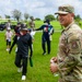 A U.S. Air Force service member in OCP (Operational Camouflage Pattern) uniform stands smiling on a grassy field while a group of children and adults gather behind him. The patch on his shoulder indicates affiliation with the U.S. Air Force. The setting appears to be an outdoor community or educational event, possibly on or near a military installation.