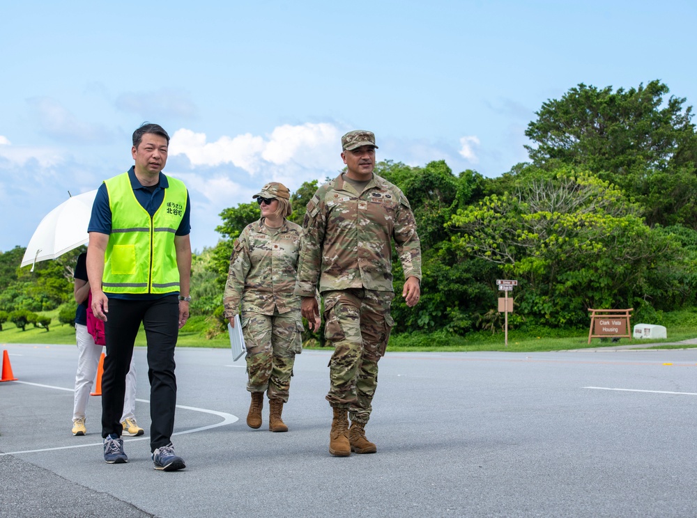 Three individuals are walking together on a paved road or tarmac in what appears to be a military installation. Two individuals are wearing US military camouflage uniforms (OCP pattern), while a third person wears a bright yellow safety vest with Japanese text and carries a white umbrella. A sign reading 'Clark Vista Housing' is visible in the background, suggesting a US military base, likely in Japan or Okinawa.