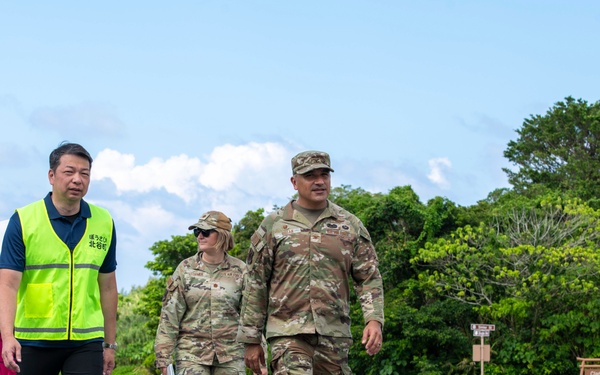 Three individuals are walking together on a paved road or tarmac in what appears to be a military installation. Two individuals are wearing US military camouflage uniforms (OCP pattern), while a third person wears a bright yellow safety vest with Japanese text and carries a white umbrella. A sign reading 'Clark Vista Housing' is visible in the background, suggesting a US military base, likely in Japan or Okinawa.