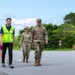 Three individuals are walking together on a paved road or tarmac in what appears to be a military installation. Two individuals are wearing US military camouflage uniforms (OCP pattern), while a third person wears a bright yellow safety vest with Japanese text and carries a white umbrella. A sign reading 'Clark Vista Housing' is visible in the background, suggesting a US military base, likely in Japan or Okinawa.