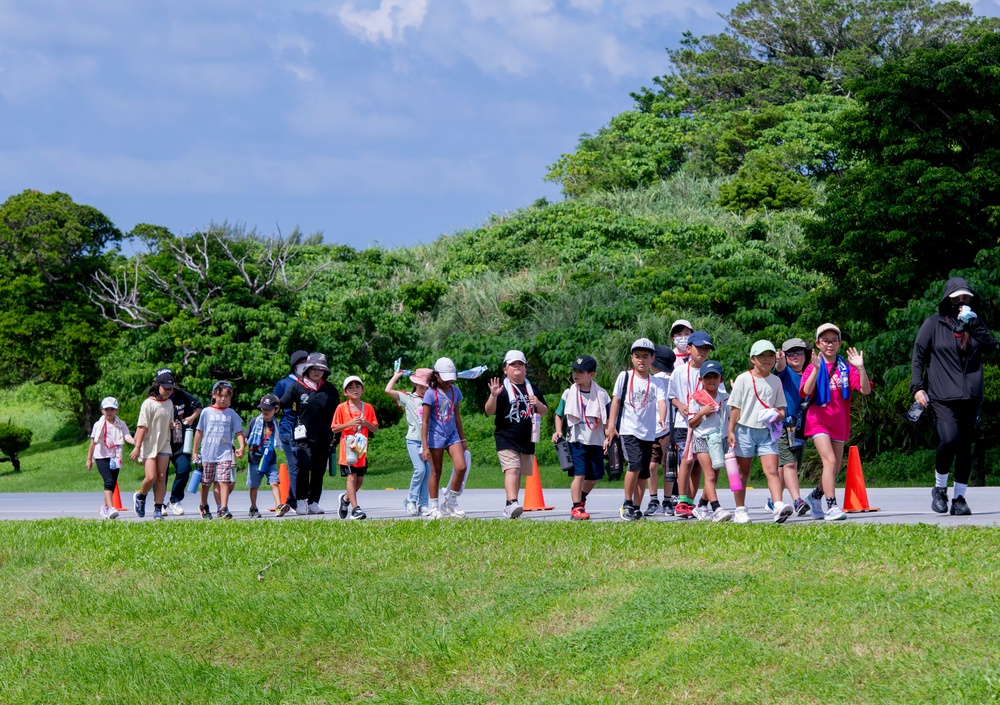 A group of young children, likely on a school field trip, walk in a line along a paved path flanked by orange traffic cones. They are accompanied by several adult supervisors and are dressed in casual summer clothing with hats and lanyards. The setting appears to be an outdoor park or recreational area with lush green trees and grass under a partly cloudy sky.