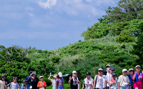 A group of young children, likely on a school field trip, walk in a line along a paved path flanked by orange traffic cones. They are accompanied by several adult supervisors and are dressed in casual summer clothing with hats and lanyards. The setting appears to be an outdoor park or recreational area with lush green trees and grass under a partly cloudy sky.