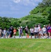 A group of young children, likely on a school field trip, walk in a line along a paved path flanked by orange traffic cones. They are accompanied by several adult supervisors and are dressed in casual summer clothing with hats and lanyards. The setting appears to be an outdoor park or recreational area with lush green trees and grass under a partly cloudy sky.