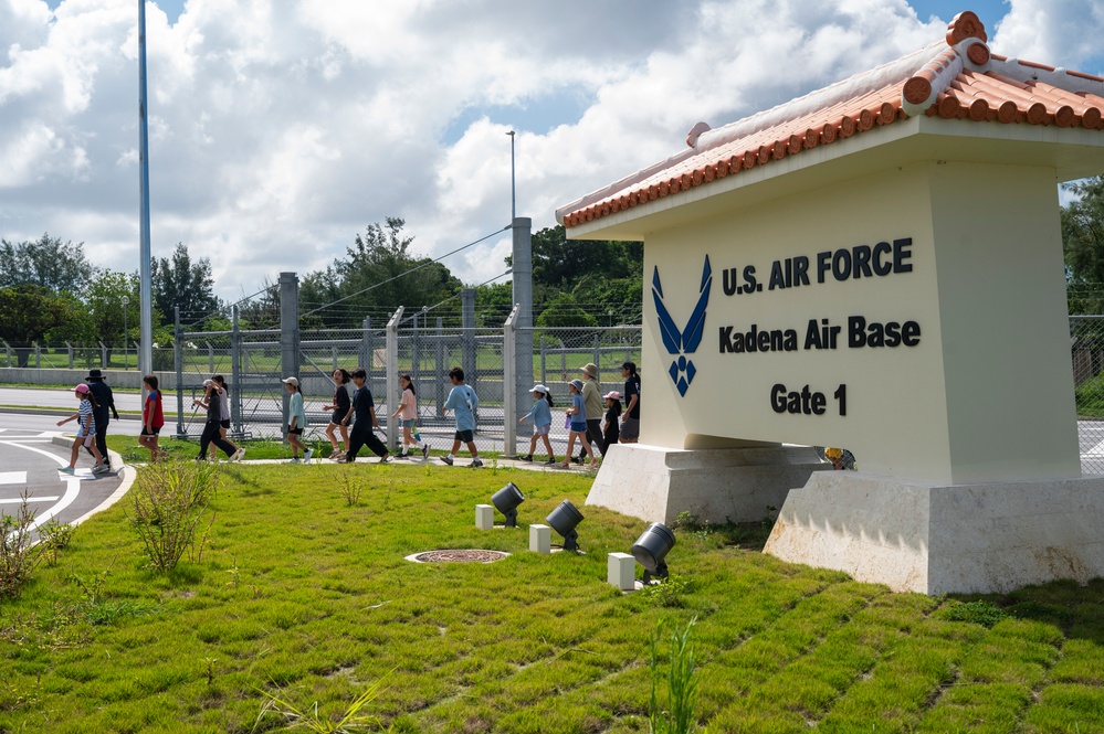 A group of children and adults walk past the Gate 1 entrance sign of Kadena Air Base, a U.S. Air Force installation. The sign features the U.S. Air Force logo and is topped with a traditional Okinawan-style red tile roof, reflecting the base's location in Okinawa, Japan. The group appears to be on a guided tour or organized visit to the base.