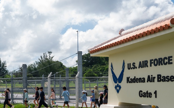 A group of children and adults walk past the Gate 1 entrance sign of Kadena Air Base, a U.S. Air Force installation. The sign features the U.S. Air Force logo and is topped with a traditional Okinawan-style red tile roof, reflecting the base's location in Okinawa, Japan. The group appears to be on a guided tour or organized visit to the base.