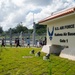 A group of children and adults walk past the Gate 1 entrance sign of Kadena Air Base, a U.S. Air Force installation. The sign features the U.S. Air Force logo and is topped with a traditional Okinawan-style red tile roof, reflecting the base's location in Okinawa, Japan. The group appears to be on a guided tour or organized visit to the base.