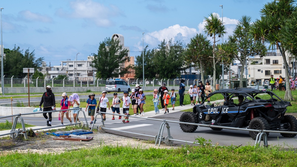 A group of children accompanied by adult chaperones crosses a road at a marked crosswalk, likely during a school field trip or organized event. A black police-marked off-road utility vehicle (UTV) labeled 'POLICE' with the number '5' is parked nearby. The setting appears to be a military base or secured facility, suggested by perimeter fencing and the style of infrastructure.