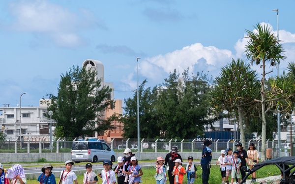 A group of children accompanied by adult chaperones crosses a road at a marked crosswalk, likely during a school field trip or organized event. A black police-marked off-road utility vehicle (UTV) labeled 'POLICE' with the number '5' is parked nearby. The setting appears to be a military base or secured facility, suggested by perimeter fencing and the style of infrastructure.