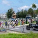 A group of children accompanied by adult chaperones crosses a road at a marked crosswalk, likely during a school field trip or organized event. A black police-marked off-road utility vehicle (UTV) labeled 'POLICE' with the number '5' is parked nearby. The setting appears to be a military base or secured facility, suggested by perimeter fencing and the style of infrastructure.