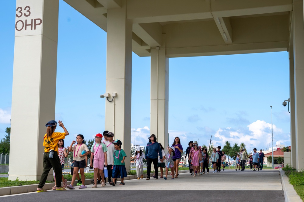 A group of children accompanied by adult chaperones walks in a line under a large covered structure or overpass marked '33 OHP'. The scene appears to be a school field trip or organized group outing in what looks like a facility or campus environment. A guide or leader at the front is gesturing with a raised hand, directing the group.