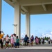 A group of children accompanied by adult chaperones walks in a line under a large covered structure or overpass marked '33 OHP'. The scene appears to be a school field trip or organized group outing in what looks like a facility or campus environment. A guide or leader at the front is gesturing with a raised hand, directing the group.