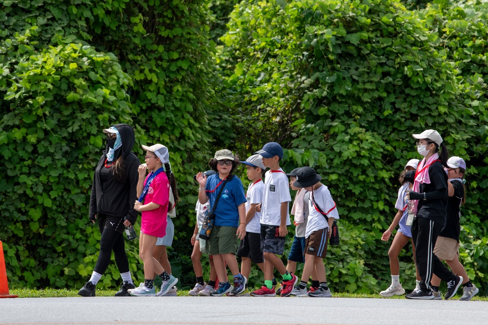 A group of children, approximately 8-10 in number, are walking in a line along a paved path outdoors, accompanied by at least two adult chaperones. The setting appears to be a park or outdoor educational venue, with dense green foliage in the background. Several individuals are wearing face masks and hats, suggesting sun protection measures and possible COVID-era health precautions.