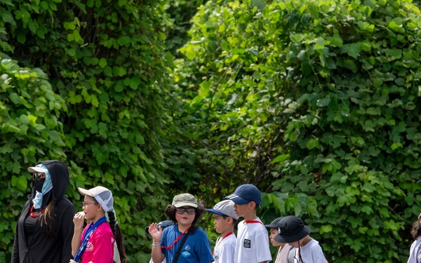 A group of children, approximately 8-10 in number, are walking in a line along a paved path outdoors, accompanied by at least two adult chaperones. The setting appears to be a park or outdoor educational venue, with dense green foliage in the background. Several individuals are wearing face masks and hats, suggesting sun protection measures and possible COVID-era health precautions.