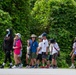 A group of children, approximately 8-10 in number, are walking in a line along a paved path outdoors, accompanied by at least two adult chaperones. The setting appears to be a park or outdoor educational venue, with dense green foliage in the background. Several individuals are wearing face masks and hats, suggesting sun protection measures and possible COVID-era health precautions.
