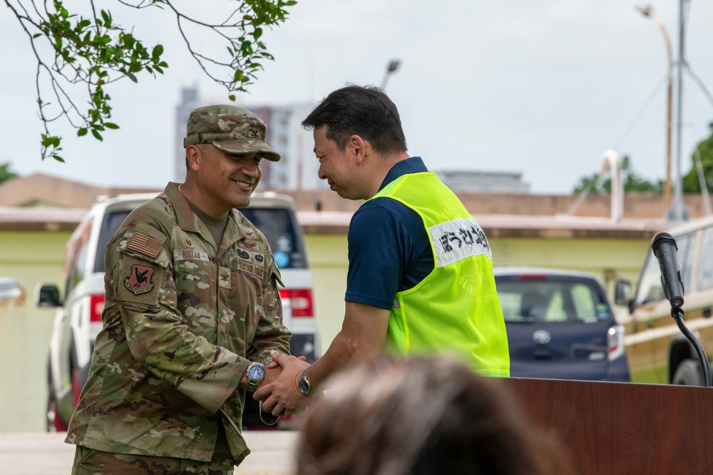 A U.S. Air Force servicemember in OCP (Operational Camouflage Pattern) uniform is shaking hands with a civilian wearing a high-visibility yellow vest bearing Japanese characters. The interaction appears to be a formal or ceremonial greeting, likely at a joint U.S.-Japanese civil-military cooperation event. The setting appears to be an outdoor area near a parking lot, possibly on or near a U.S. military installation in Japan.