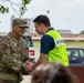 A U.S. Air Force servicemember in OCP (Operational Camouflage Pattern) uniform is shaking hands with a civilian wearing a high-visibility yellow vest bearing Japanese characters. The interaction appears to be a formal or ceremonial greeting, likely at a joint U.S.-Japanese civil-military cooperation event. The setting appears to be an outdoor area near a parking lot, possibly on or near a U.S. military installation in Japan.