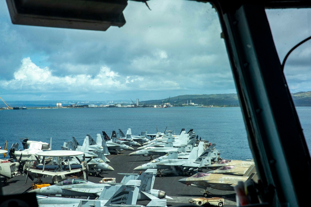 The image is taken from the island/bridge of a US Navy aircraft carrier, looking down across a densely packed flight deck filled with multiple fixed-wing aircraft. The carrier appears to be anchored or moored near a port facility, with a coastline, industrial infrastructure, and green hills visible in the background across the water. The perspective suggests the photo was taken through a bridge window.