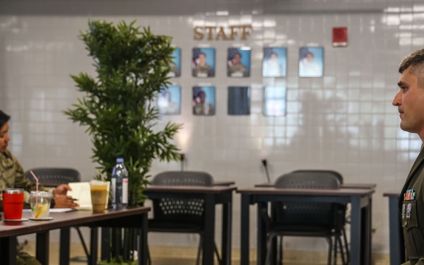 A U.S. Army staff sergeant in dress green uniform sits facing a panel of two soldiers in combat uniforms at a table, suggesting a formal board or interview setting. The room appears to be a military administrative or meeting space with a 'STAFF' sign and portrait photos on the wall. The seated soldier's posture and attire indicate this is likely a promotion board or formal evaluation.