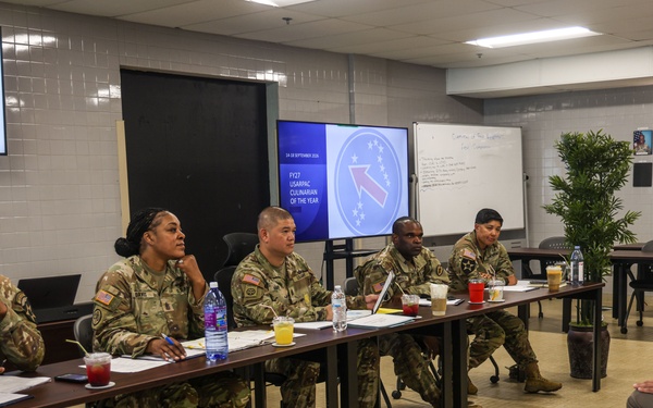A panel of five U.S. Army soldiers in camouflage uniforms sits at a long table facing a single soldier in dress uniform, suggesting a formal board or evaluation interview. A presentation screen in the background displays 'FY27 USARPAC Culinary of the Year,' indicating this is a military culinary competition board. The setting appears to be a military dining facility or conference room with a 'STAFF' sign and personnel photos visible on the wall.