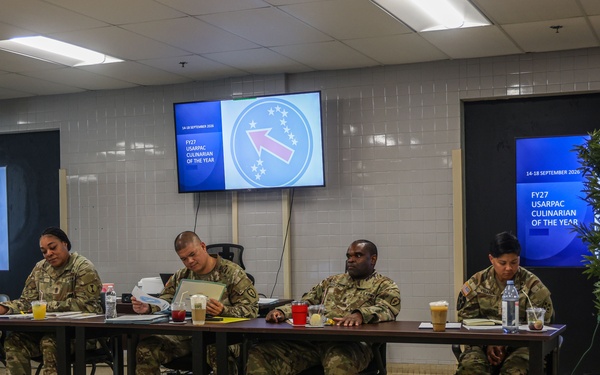 A panel of U.S. Army soldiers in camouflage uniforms are seated at a long table, appearing to serve as judges or evaluators for the 'FY27 USARPAC Culinarian of the Year' competition, held 14-18 September. Multiple display screens showing the competition branding are visible on the walls. Two additional individuals, one in dress uniform and one in civilian-style attire, are seated separately at smaller tables.
