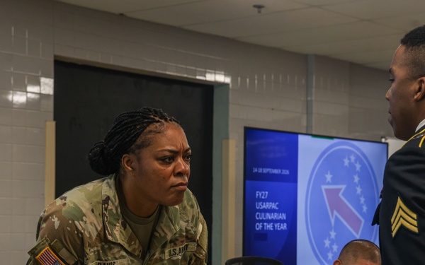 A U.S. Army briefing or competition event related to the 'FY27 USARPAC Culinarian of the Year' is taking place in a conference room. A female soldier identified by nametape as 'Davis' leans over a table in conversation with a male soldier in Army Service Uniform (ASU) dress blues bearing Sergeant chevrons. Two additional soldiers, including one identified as 'Crawford,' are seated at the table reviewing documents.