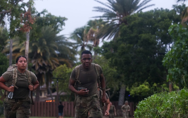 Two military personnel in camouflage uniforms and tactical backpacks are running or fast-marching along a paved path in a tropical setting. Additional uniformed personnel are visible in the background, suggesting a group physical training or ruck march exercise. The environment features palm trees and lush vegetation consistent with a tropical military installation.