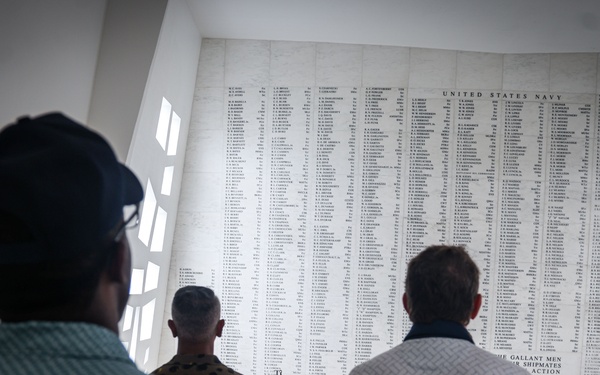Several visitors stand facing a large marble memorial wall inscribed with names under the heading 'United States Navy.' The setting appears to be the USS Arizona Memorial at Pearl Harbor, Hawaii, based on the visible inscription referencing 'Arizona' and 'gallant men' and 'shipmates.' One individual appears to be in military camouflage uniform, and a red floral lei is visible, consistent with Hawaiian memorial traditions.