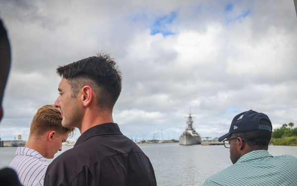 Several people are standing on what appears to be an observation deck or memorial platform, looking out over a harbor toward a large naval warship in the distance. The setting, with its white structure and view of a battleship, is consistent with the USS Arizona Memorial or a similar naval heritage site at Pearl Harbor, Hawaii. The group appears to be visitors observing the historic vessel across the water.