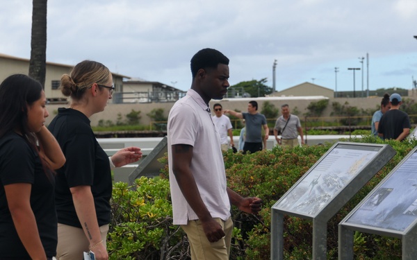 Three individuals in the foreground are examining outdoor interpretive/informational display panels along a paved walkway, likely at a historical or military site. The setting appears to be a well-maintained outdoor memorial or visitor area with landscaped shrubs and military-style buildings visible in the background. Additional visitors can be seen walking in the background.