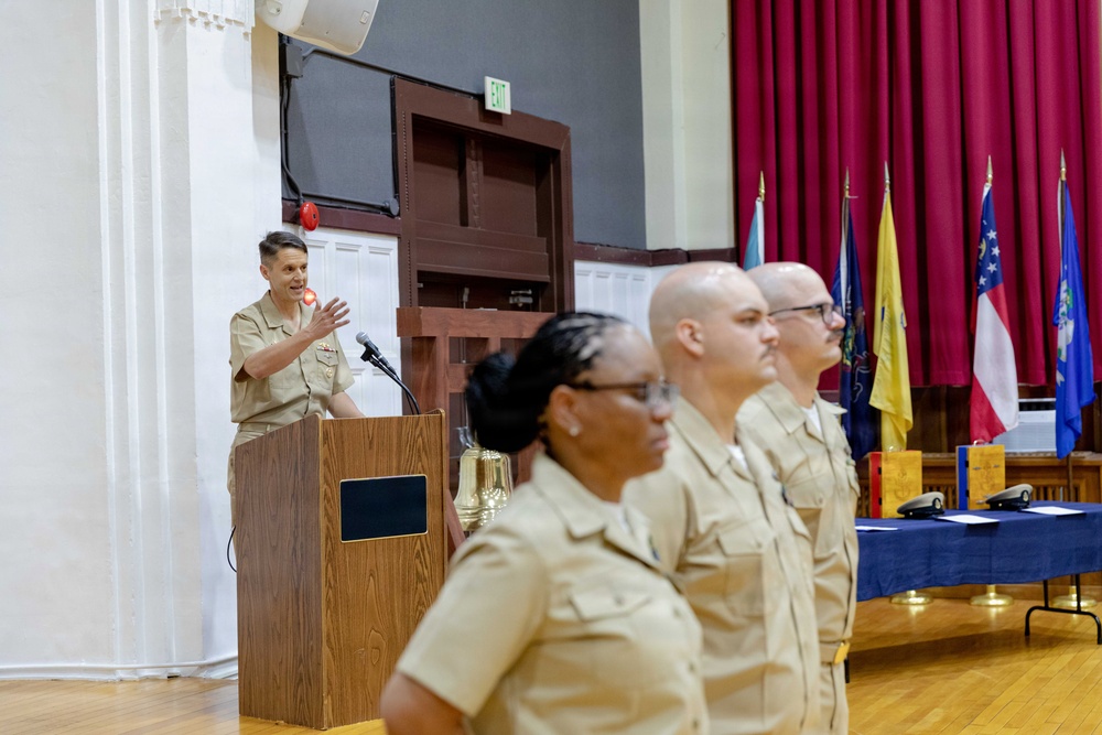 A military ceremony taking place in a formal hall, with an officer speaking at a wooden podium while three uniformed personnel stand at attention in the foreground. All individuals are wearing tan/khaki military uniforms consistent with U.S. Navy service dress. Multiple state and organizational flags are displayed in the background alongside a blue-draped ceremonial table.