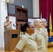 A military ceremony taking place in a formal hall, with an officer speaking at a wooden podium while three uniformed personnel stand at attention in the foreground. All individuals are wearing tan/khaki military uniforms consistent with U.S. Navy service dress. Multiple state and organizational flags are displayed in the background alongside a blue-draped ceremonial table.