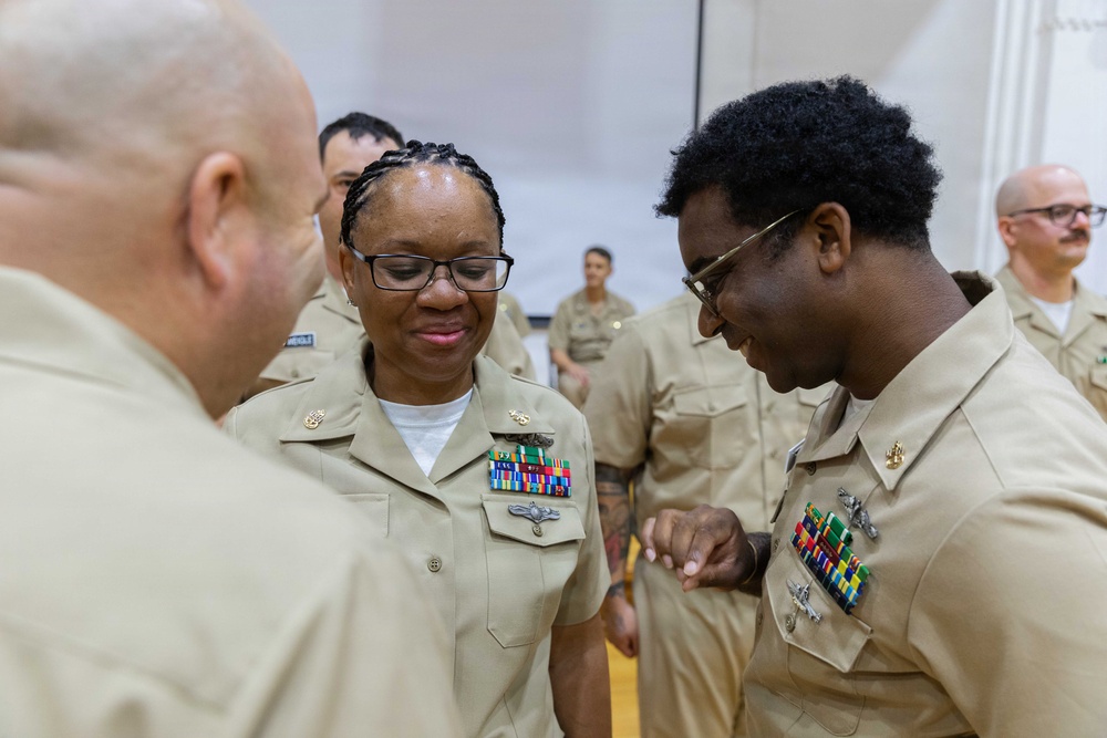 A group of U.S. Navy personnel in khaki service uniforms are gathered in what appears to be an indoor ceremony or formal event. A central female sailor with glasses and multiple service ribbons and badges is being attended to by another sailor, possibly having an insignia or pin affixed to her uniform. The setting suggests a promotion, award, or pinning ceremony.