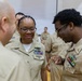 A group of U.S. Navy personnel in khaki service uniforms are gathered in what appears to be an indoor ceremony or formal event. A central female sailor with glasses and multiple service ribbons and badges is being attended to by another sailor, possibly having an insignia or pin affixed to her uniform. The setting suggests a promotion, award, or pinning ceremony.