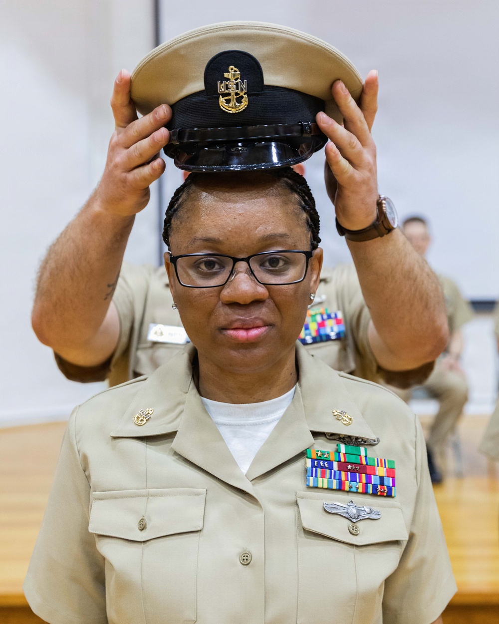 A U.S. Navy enlisted service member is having a Chief Petty Officer combination cover (cap) placed on her head by another individual, indicating a promotion or pinning ceremony. She is wearing the tan Service Uniform (SU) with multiple ribbon awards, a warfare device badge, and collar insignia. The setting appears to be an indoor ceremonial space such as a gymnasium or meeting hall.