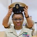 A U.S. Navy enlisted service member is having a Chief Petty Officer combination cover (cap) placed on her head by another individual, indicating a promotion or pinning ceremony. She is wearing the tan Service Uniform (SU) with multiple ribbon awards, a warfare device badge, and collar insignia. The setting appears to be an indoor ceremonial space such as a gymnasium or meeting hall.