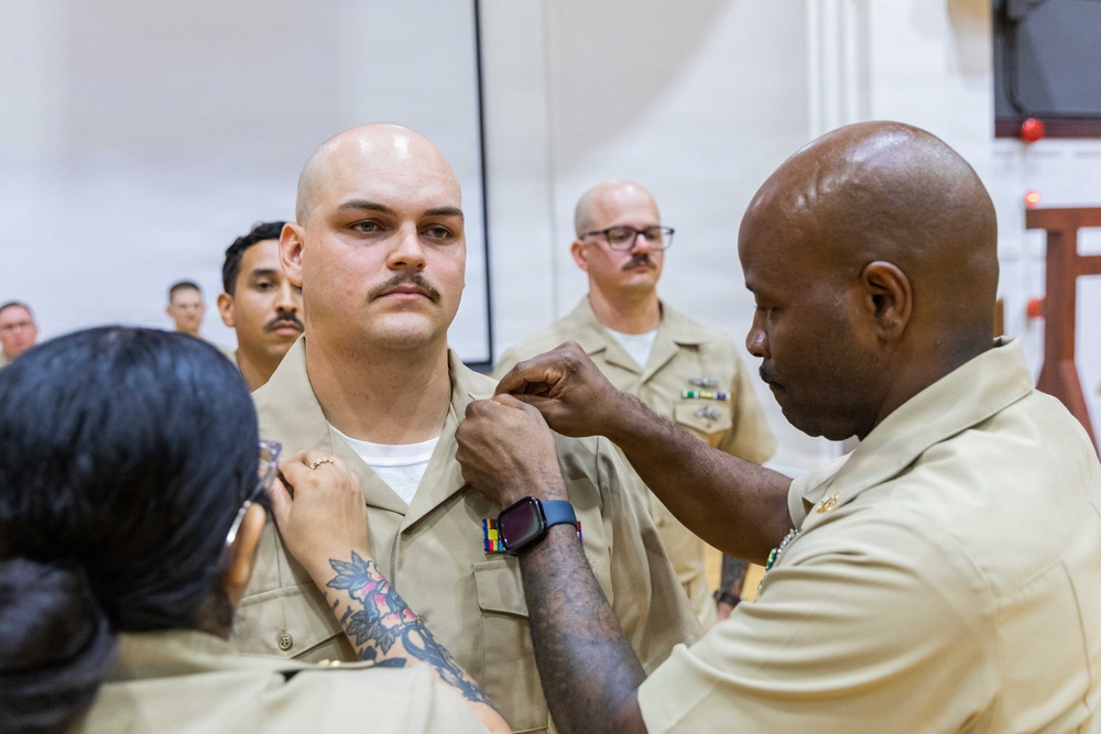 A military pinning ceremony is taking place, likely in the U.S. Navy, where two individuals are simultaneously pinning insignia or rank devices onto the collar of a bald male sailor in a khaki uniform. The recipient stands at attention with a serious expression while witnesses in matching uniforms observe in the background. This type of ceremony typically marks a promotion or qualification achievement.