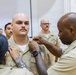 A military pinning ceremony is taking place, likely in the U.S. Navy, where two individuals are simultaneously pinning insignia or rank devices onto the collar of a bald male sailor in a khaki uniform. The recipient stands at attention with a serious expression while witnesses in matching uniforms observe in the background. This type of ceremony typically marks a promotion or qualification achievement.