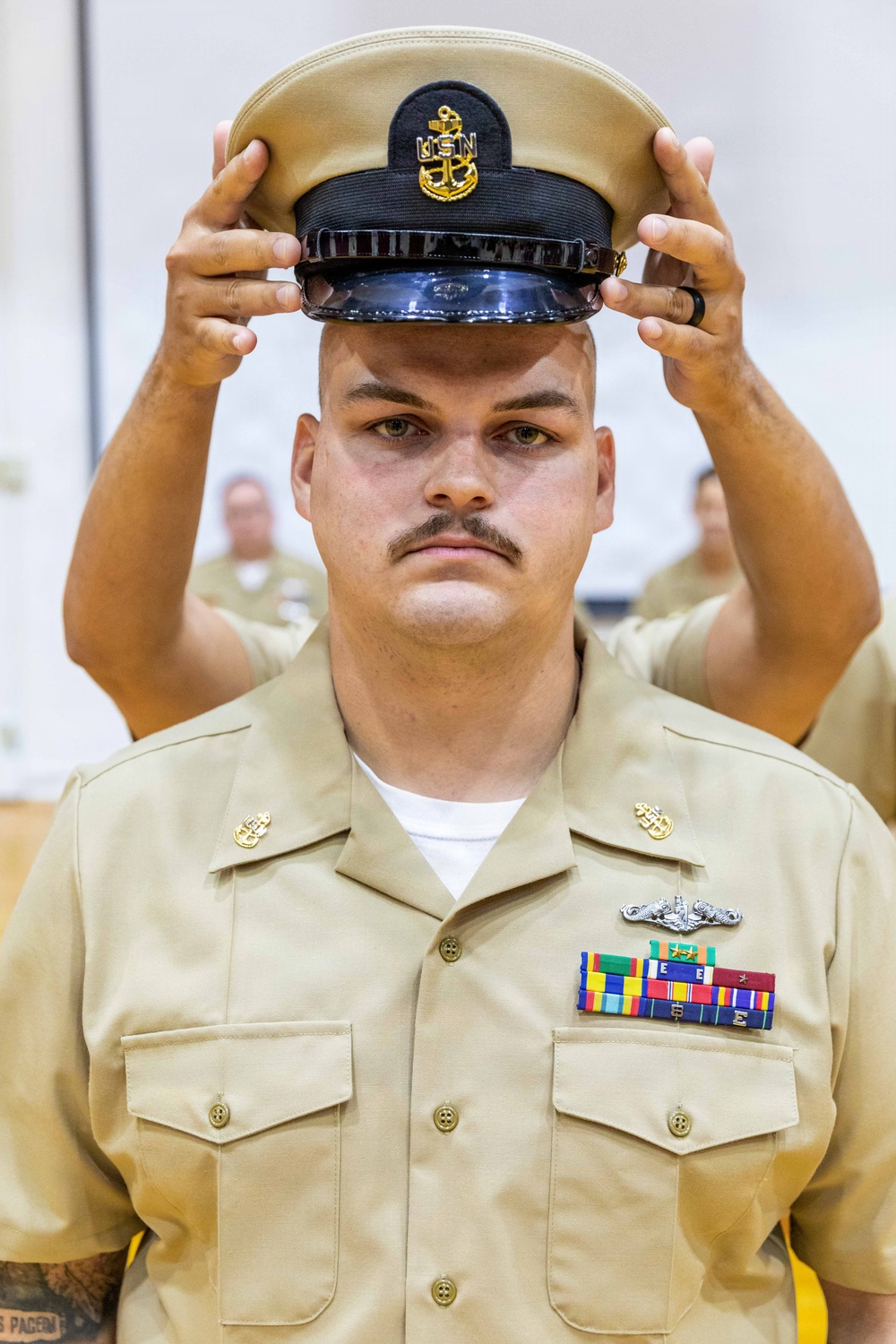 A U.S. Navy Chief Petty Officer is placing his combination cover (cap) on his head during what appears to be a chief pinning or frocking ceremony. He is wearing the khaki service uniform with a submarine warfare insignia, multiple service ribbons, and chief petty officer collar devices. The formal setting and solemn expression suggest this is a significant military advancement ceremony.