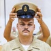 A U.S. Navy Chief Petty Officer is placing his combination cover (cap) on his head during what appears to be a chief pinning or frocking ceremony. He is wearing the khaki service uniform with a submarine warfare insignia, multiple service ribbons, and chief petty officer collar devices. The formal setting and solemn expression suggest this is a significant military advancement ceremony.