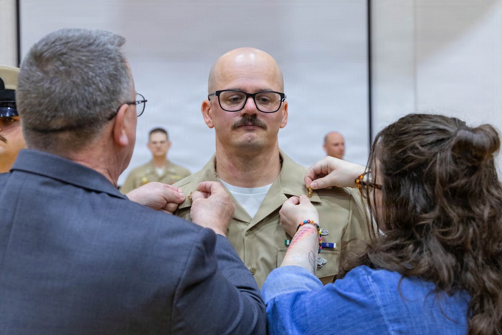 A military promotion or award ceremony is taking place indoors. A bald man wearing glasses and a tan/khaki military uniform is having insignia or rank devices pinned onto his collar by two individuals — an older man in civilian clothing on the left and a younger woman in a blue top on the right. Additional uniformed military personnel are visible in the background.