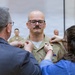 A military promotion or award ceremony is taking place indoors. A bald man wearing glasses and a tan/khaki military uniform is having insignia or rank devices pinned onto his collar by two individuals — an older man in civilian clothing on the left and a younger woman in a blue top on the right. Additional uniformed military personnel are visible in the background.